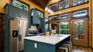 A modern kitchen with a stainless steel refrigerator, a large island with stools, and a view of the outdoors through large windows.