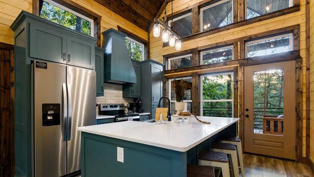A modern kitchen with a stainless steel refrigerator, a large island with stools, and a view of the outdoors through large windows.