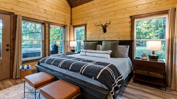 A wooden cabin bedroom with a bed, bench, nightstand, and a mounted deer head.