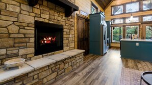 A stone fireplace with a lit fire, a kitchen area with a refrigerator, and large windows offering a view of the outdoors.