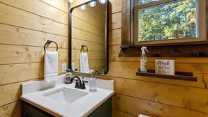 A wooden bathroom with a white sink, a mirror with lights, and a window with a view of trees.