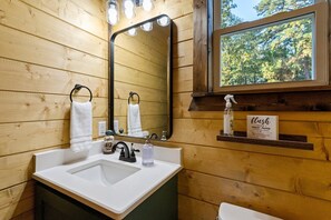 Une salle de bain en bois avec un lavabo blanc, un miroir avec éclairage intégré et une fenêtre donnant sur les arbres.
