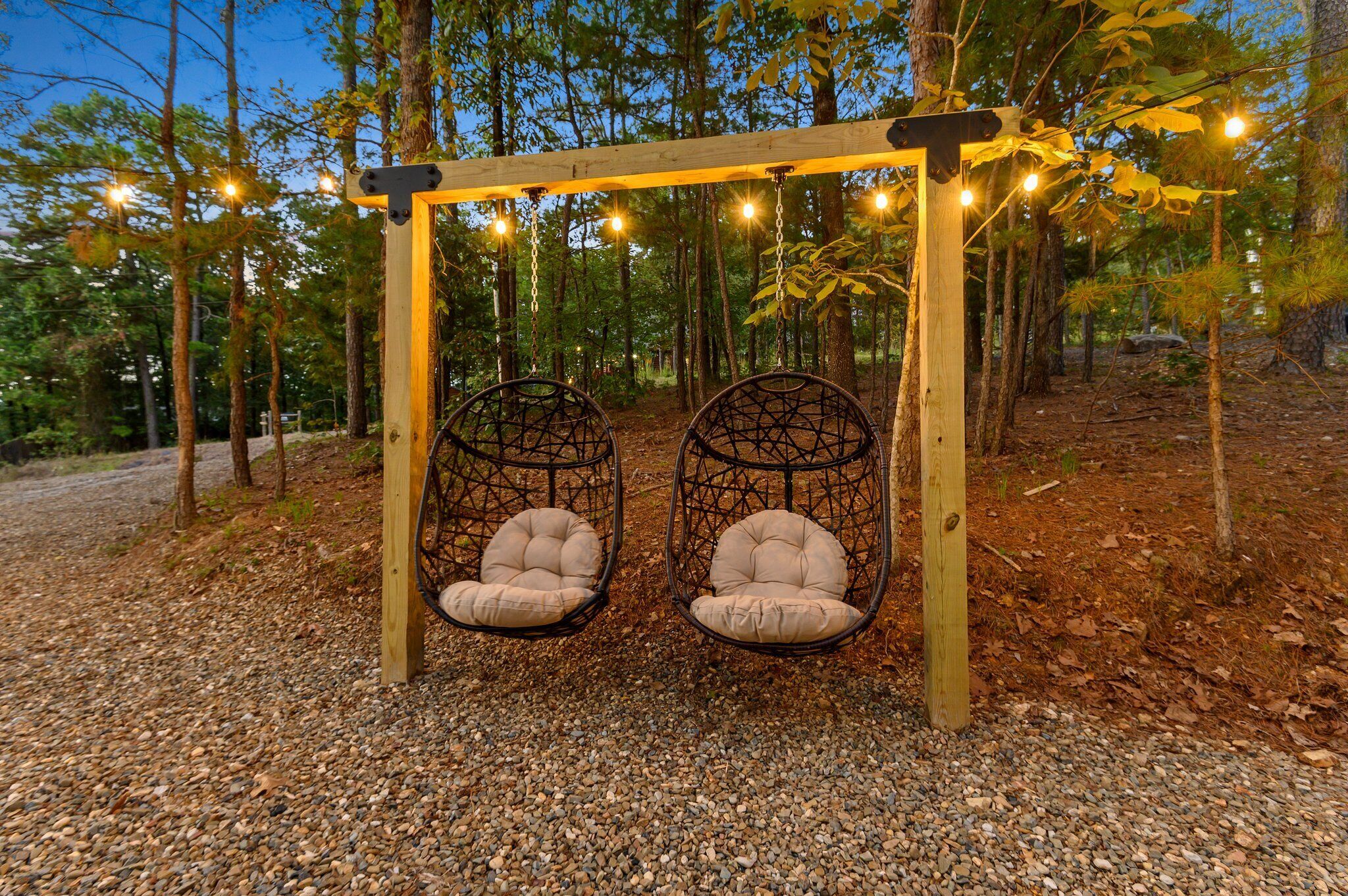 Une balançoire en bois avec deux sièges rembourrés, entourée d’une zone boisée illuminée de guirlandes lumineuses.
