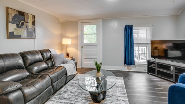 A living room with a leather sofa, a glass coffee table with a plant, a flat-screen TV, and a door leading to a balcony with a view. - Pet-Friendly Auburn Apartment w/ Balcony! (Auburn)