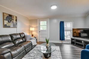 A living room with a leather sofa, a glass coffee table with a plant, a flat-screen TV, and a door leading to a balcony with a view. - Pet-Friendly Auburn Apartment w/ Balcony! (Auburn)