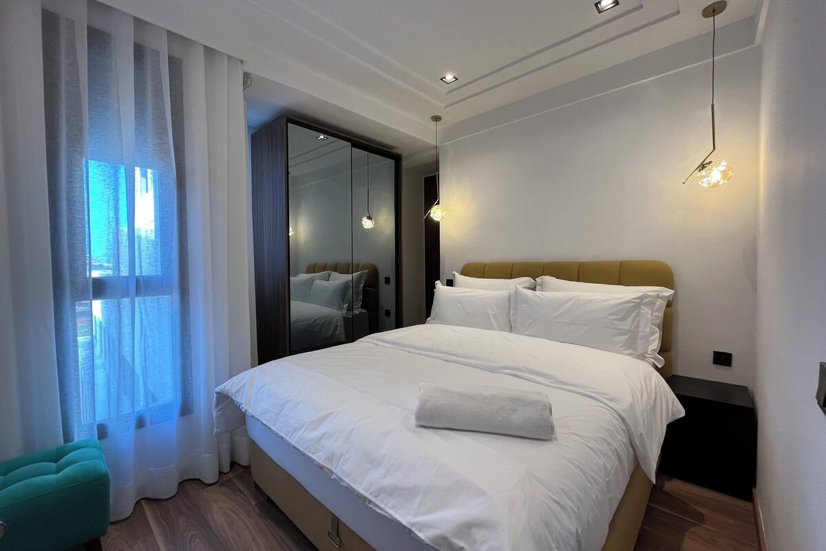 A modern hotel room with a large bed, a mirror, and a view of the city through the window.