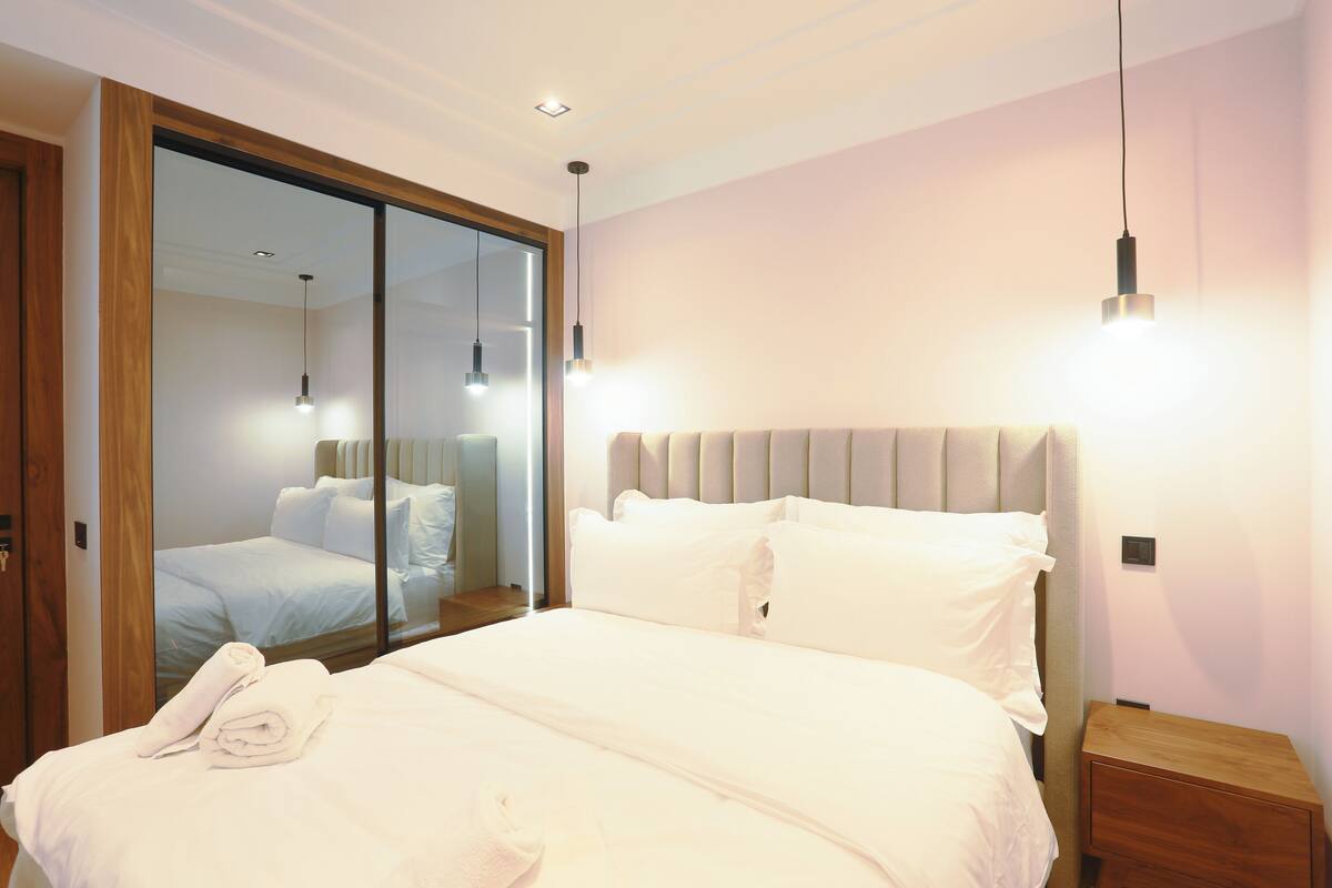 A modern hotel room with a large bed, a wooden nightstand, and a mirror reflecting the room.