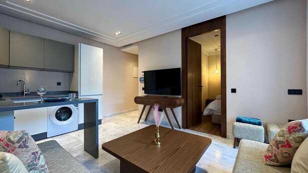 A modern living room with a sofa, a coffee table, and a TV. There is a kitchen area with a washing machine and a dining table. - Les Joyeux Luxury Suites & Appart (Casablanca)
