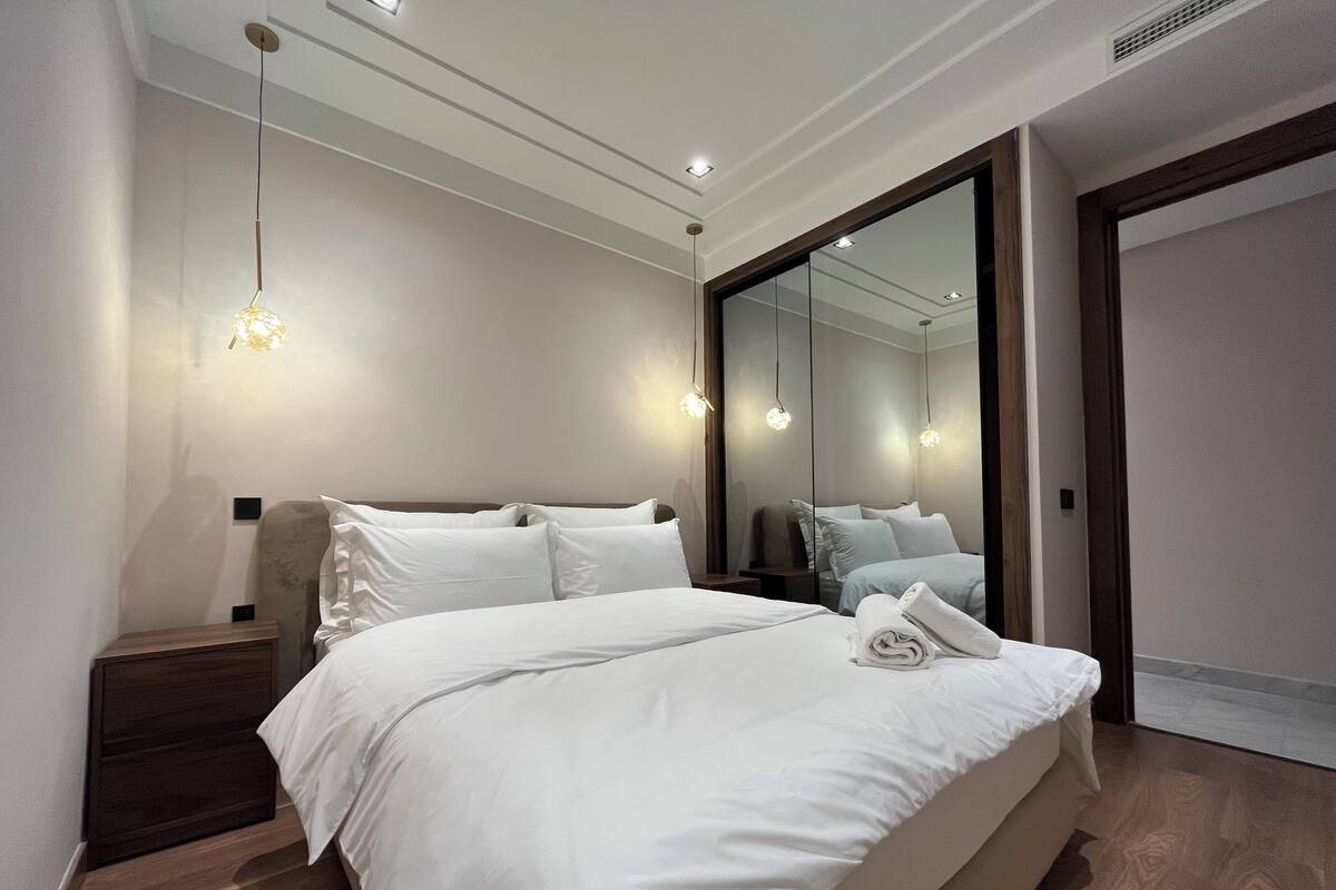 A modern bedroom with a large bed, bedside tables, a mirror, and recessed ceiling lighting.