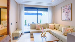 A modern living room with a sofa, ottoman, and coffee table. There is a large window with curtains, a painting on the wall, and a balcony with plants. - Les Joyeux Luxury Suites & Appart (Casablanca)