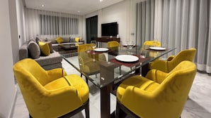 A modern living room with a glass dining table, yellow chairs, a grey sofa with yellow cushions, and a flat-screen TV on the wall. - Les Joyeux Luxury Suites & Appart (Casablanca)