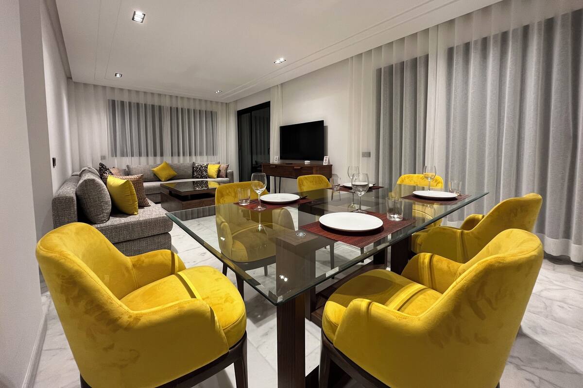 A modern living room with a glass dining table, yellow chairs, a grey sofa with yellow cushions, and a flat-screen TV on the wall.