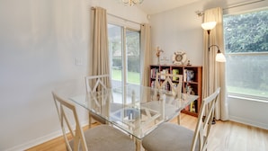 Un coin repas avec une table en verre, des chaises blanches, un lustre, une bibliothèque et une vue sur la verdure à l’extérieur.