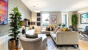 Ein modernes Wohnzimmer mit Sofa, Sessel und Couchtisch. Ein großes Fenster mit Vorhängen, eine Pflanze und Kunstwerke an den Wänden.