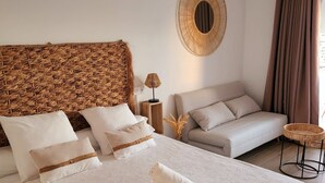 Ein ordentlich bezogenes Bett mit gerolltem Handtuch, einem Weiden-Kopfstück, einem runden Wandbild, einem Nachttisch mit Lampe und einem Sofa in einem modernen, hellen Raum.