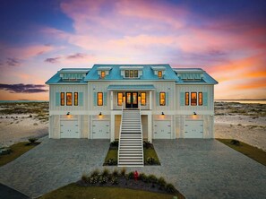 A large, two-story beachfront house with a prominent front entrance, multiple garage doors, and a driveway leading to the entrance.