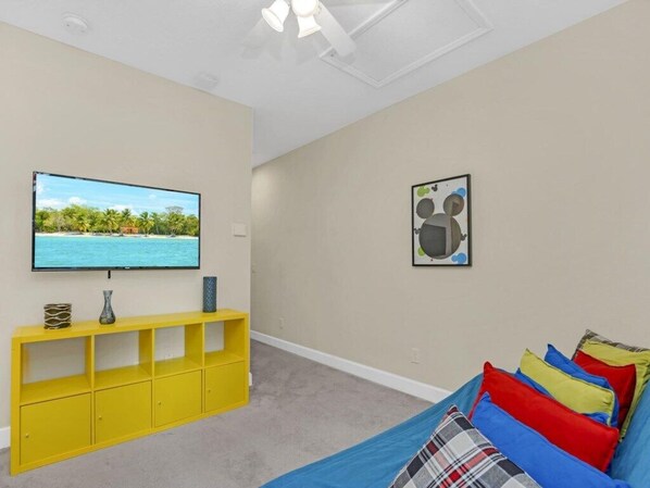 Una habitación con televisor en la pared, un mueble de entretenimiento amarillo, una cama con almohadones de colores y un cuadro enmarcado en la pared.
