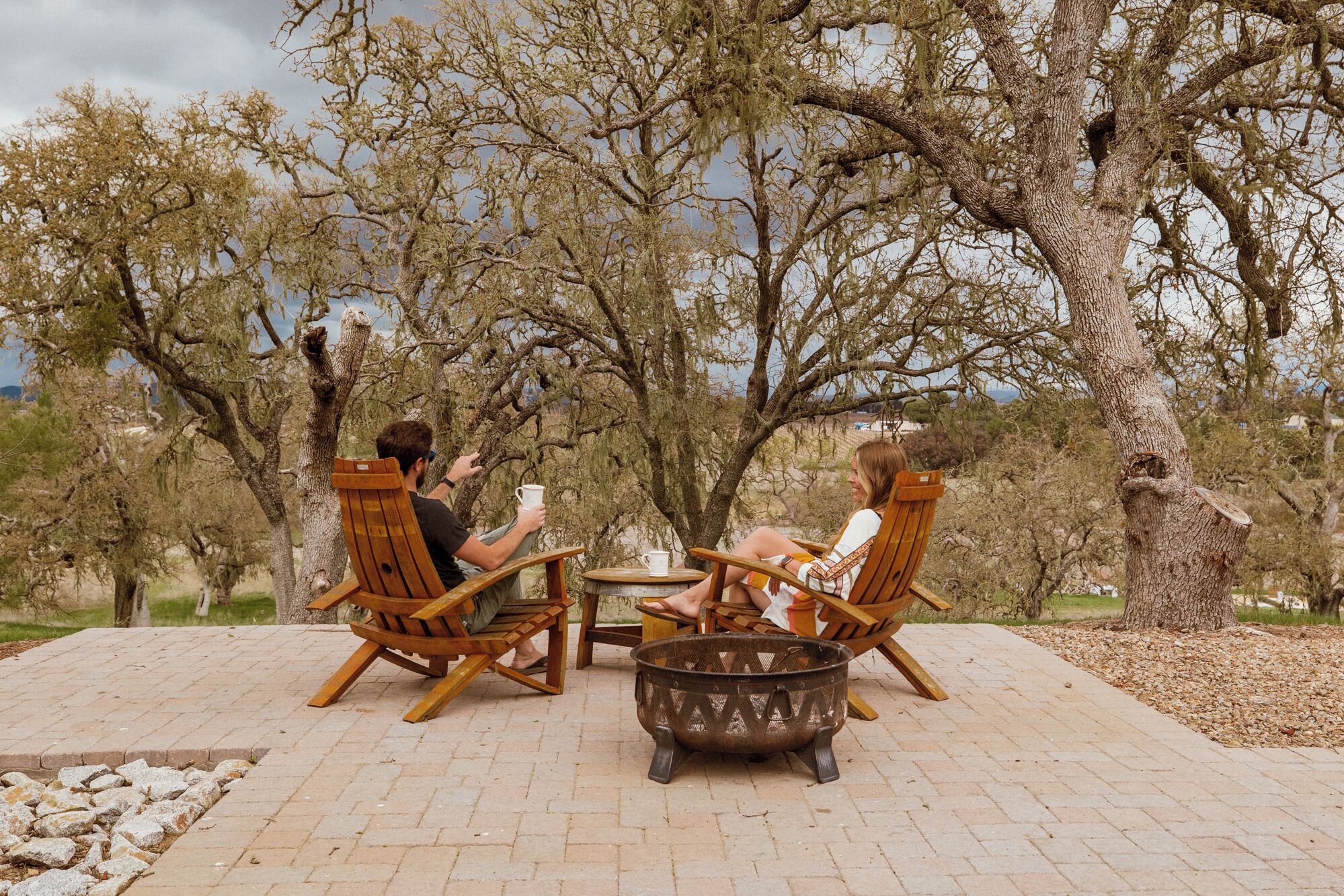 Two people seated at a table with a fire pit in between, surrounded by trees.