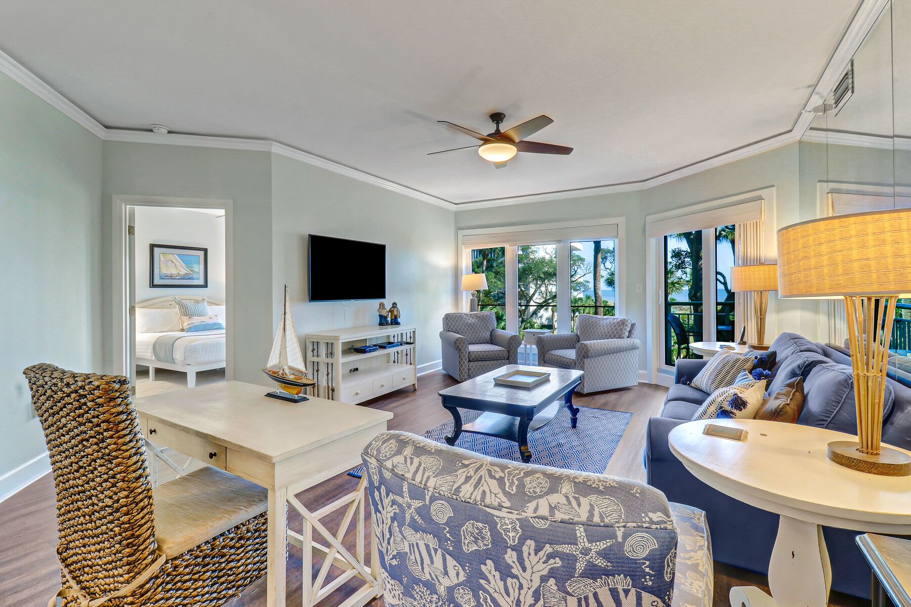 A spacious living room with a ceiling fan, a TV mounted on the wall, and a view of the outdoors through large windows.
