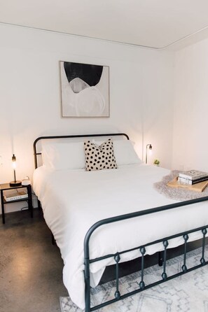 A neatly made bed with white bedding, a black metal frame, and a single polka-dotted pillow. There is a bedside table with a lamp and a book. A framed abstract artwork hangs above the bed.