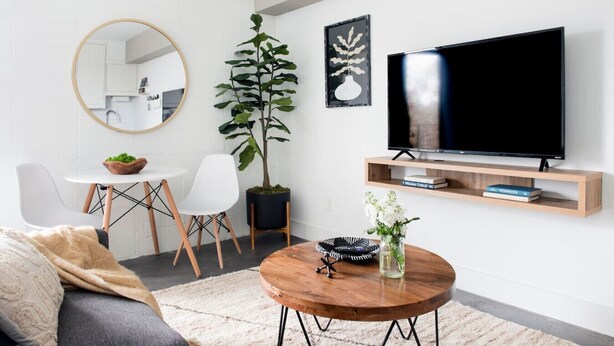 A modern living room with a round wooden coffee table, a sofa, a dining table with chairs, a large potted plant, a flat-screen TV on a wooden stand, and a mirror on the wall. - La Penelope – Walk to Ponce City Market + Rooftop (Atlanta)