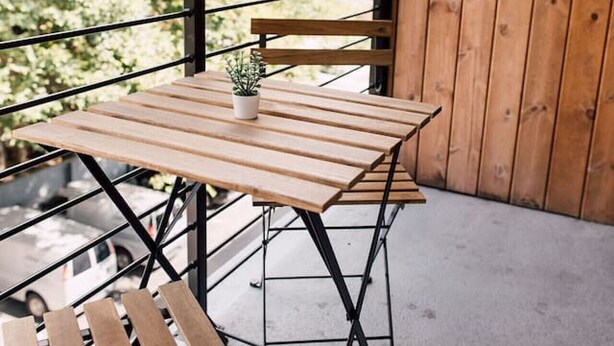 Un conjunto de mesa y banco de madera en un balcón con barandilla de metal, una planta en maceta y vistas a árboles.