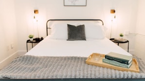 A neatly made bed with a gray pillow, a bedside table with a lamp, and a tray with books and a glass. - The Eleanor – Rooftop + City Skyline Views ATL (Atlanta)