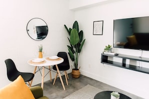 A modern living room with a round dining table, black chairs, a large potted plant, a flat-screen TV, and a mounted mirror.