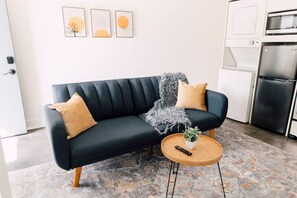 A modern living room with a dark blue sofa, two beige cushions, a gray throw blanket, and a small wooden coffee table with a plant and remote control.