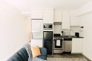 A compact kitchen with a built-in oven, microwave, and dishwasher, a gray couch with a throw blanket, and white cabinetry.