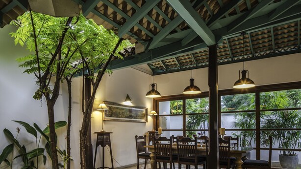 Een eetruimte met houten stoelen en tafels, een plafond met zichtbare houten balken en uitzicht op groen buiten.