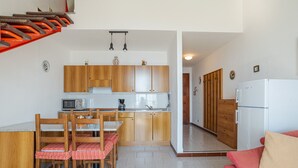Une cuisine compacte avec des meubles en bois, un four à micro-ondes et un réfrigérateur. On y trouve une table à manger avec des chaises et un coussin rouge. Un canapé rouge est visible dans l’espace salon.
