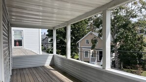 Eine überdachte Veranda mit Holzdeck, weißen Geländern und großen Fenstern, die einen Blick auf benachbarte Häuser und Bäume bietet.
