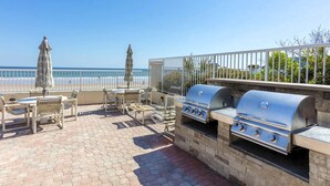 Eine Terrasse im Freien mit Grill, Sitzgelegenheiten und Sonnenschirmen mit Blick auf den Strand.