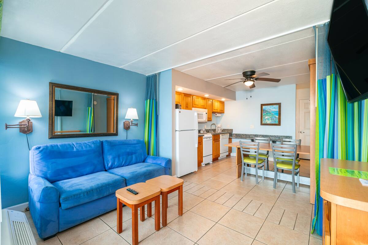 A compact living space with a blue sofa, wooden table, and chairs, a kitchenette with a refrigerator and microwave, and a ceiling fan.