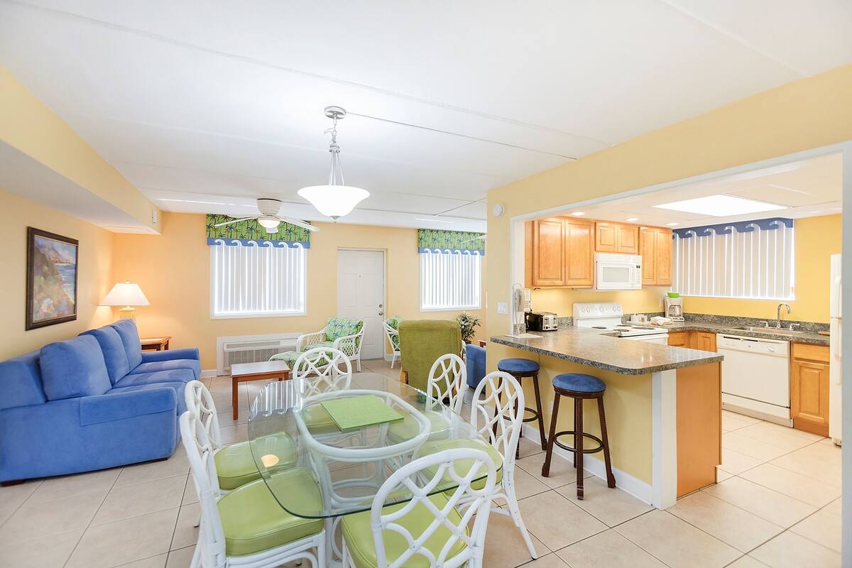 A bright living room with a blue sofa, a glass dining table with green chairs, and a kitchen area with wooden cabinets and a white refrigerator.