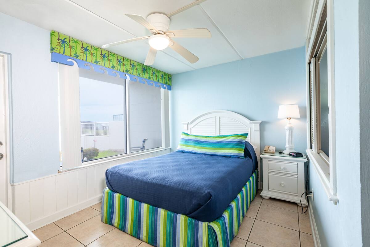 A bedroom with a bed, a nightstand, a lamp, a ceiling fan, and a window with a view.