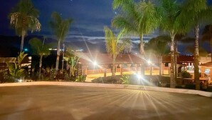 A nighttime view of a resort with palm trees, a covered area with lights, and a building in the background. - Cozy Cabin with Terrace and Panoramic View- Access to Pool (Ejido El Porvenir (Guadalupe))