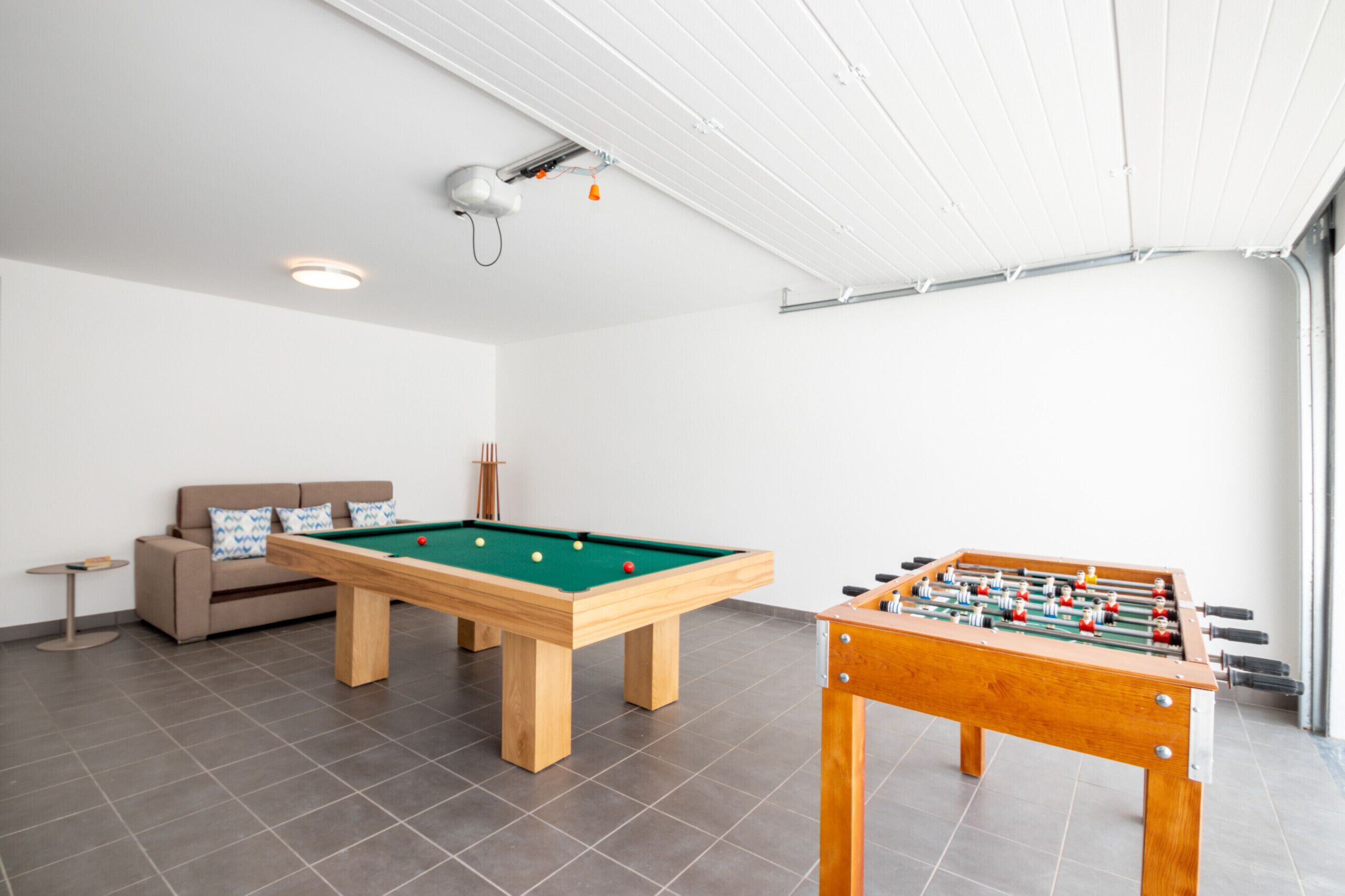 Ein Billardtisch, ein Kicker und ein Sofa in einem Raum mit hoher Decke und Fliesenboden.
