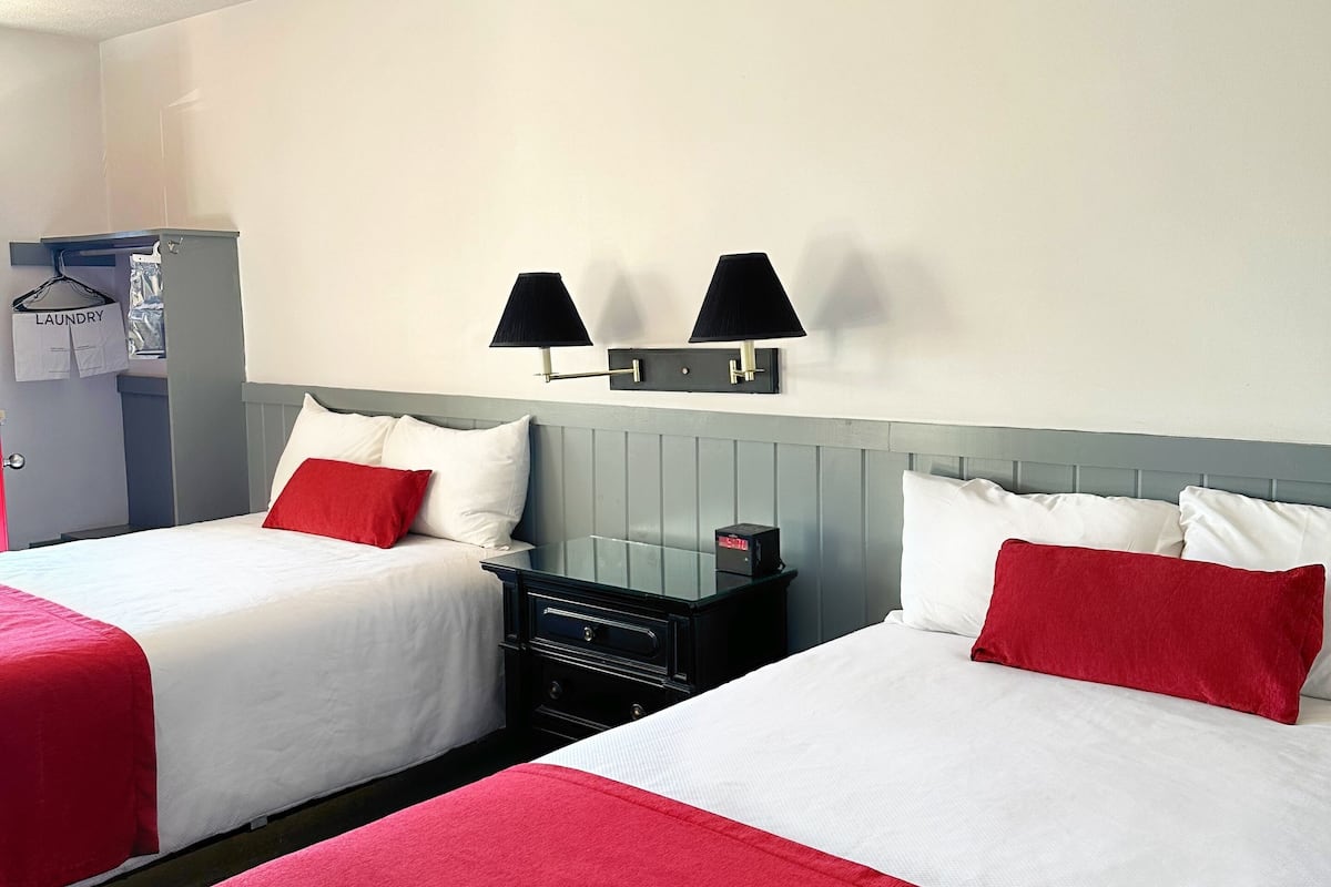 A hotel room with two beds, each with white and red bedding, a small table with a lamp, and a wardrobe.