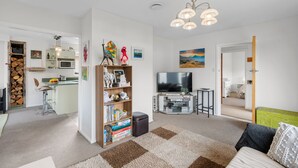 A modern living room with a sofa, a coffee table, a bookshelf, a TV, and a kitchen area in the background. - Pops Place - Omarama Holiday Home (Omarama)