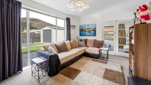 A modern living room with a sectional sofa, a small table, and a bookshelf. There is a large window offering a view of a backyard and a hill. - Pops Place - Omarama Holiday Home (Omarama)