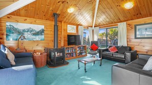 A cozy cabin interior with a fireplace, bookshelves, a television, and a large window offering a view of the outdoors.