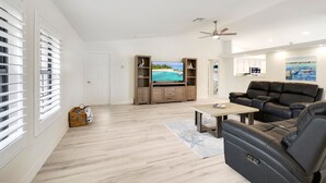 Een ruime woonkamer met een grote flatscreen-tv, een zwarte leren bank, houten kasten en een plafondventilator.