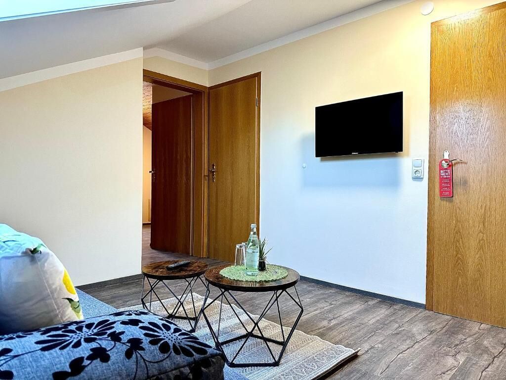 A hotel room with a bed, a small round table with a bottle and glass, a TV mounted on the wall, and a wooden door.