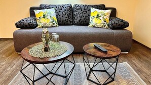 Uma sala de estar aconchegante com um sofá cinza, almofadas florais, duas mesas redondas com estrutura de metal e uma pequena planta em uma das mesas.