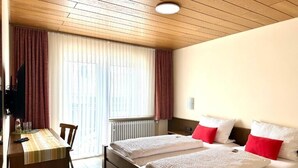 Quarto de hotel com teto de madeira, cama com roupa de cama branca e almofadas vermelhas, escrivaninha com toalha de mesa listrada e janela com cortinas.
