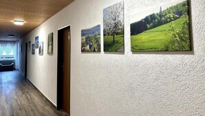 Corredor com piso de madeira e parede branca com textura. Quatro quadros emoldurados retratam paisagens e há uma porta.