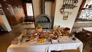 Uma mesa de jantar posta com diversos alimentos e utensílios, uma lareira e um interior rústico com soalhos de madeira e paredes de tijolo.