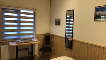A room with a bed, a radiator, a sewing machine, a window with blinds, a chair, a mirror, and a framed picture on the wall.